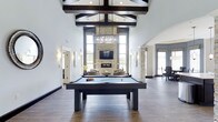 This single-floor amenity space delivers an impressive blend of modern sophistication and welcoming comfort. The centerpiece of the main room is a full-size billiards table set beneath a dramatic vaulted ceiling adorned with bold, dark exposed beams that draw the eye upward. A large crystal chandelier hangs overhead, adding a refined elegance to the expansive open layout.

Anchoring the far end of the space is a floor-to-ceiling stacked stone fireplace flanked by tall, narrow windows that invite abundant natural light and provide views of the surrounding landscape. A wall-mounted flat-screen television above the fireplace makes this lounge area equally suited for relaxed gatherings and entertainment. Comfortable seating is arranged around the hearth, creating an inviting retreat within the larger space.

The flooring throughout features rich, dark hardwood, complementing the contrast of crisp white walls and dark architectural accents. Stone-clad columns add structural character while reinforcing the upscale aesthetic. Wall sconces provide warm ambient lighting along the perimeter, enhancing the overall atmosphere. Additional rooms are accessible from the main area, suggesting versatile uses such as dining or meeting spaces. The overall ambiance is open, airy, and thoughtfully designed for both social interaction and leisure.