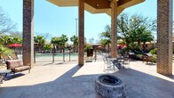 The Pool Courtyard is a well-appointed outdoor amenity space centered around a fenced swimming pool surrounded by mature palm trees and lush landscaping. A substantial covered pavilion anchored by brick columns provides shaded gathering space, featuring a built-in stone fire pit as a focal point for social gatherings. The open-air patio extends generously beyond the pavilion, offering stamped concrete flooring throughout. Outdoor dining tables and chairs are positioned poolside beneath red market umbrellas, while a grill station adds convenient outdoor cooking capability.

Adjacent to the courtyard, the fitness center is a bright, well-organized space designed for comprehensive training. Floor-to-ceiling mirrors line the walls, visually expanding the room while large windows and glass doors invite abundant natural light and direct views of the pool area. The facility is equipped with multiple treadmills, elliptical machines, and stationary bikes alongside a full cable and multi-station weight machine. A dedicated free weights section features an organized dumbbell rack, adjustable bench, and a medicine ball station. Wooden open shelving displays rolled towels and greenery, adding a polished touch. Recessed lighting, ceiling fans, and mounted televisions complete the comfortable, well-maintained environment.