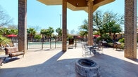 The Pool Courtyard is a well-appointed outdoor amenity space centered around a fenced swimming pool surrounded by mature palm trees and lush landscaping. A substantial covered pavilion anchored by brick columns provides shaded gathering space, featuring a built-in stone fire pit as a focal point for social gatherings. The open-air patio extends generously beyond the pavilion, offering stamped concrete flooring throughout. Outdoor dining tables and chairs are positioned poolside beneath red market umbrellas, while a grill station adds convenient outdoor cooking capability.

Adjacent to the courtyard, the fitness center is a bright, well-organized space designed for comprehensive training. Floor-to-ceiling mirrors line the walls, visually expanding the room while large windows and glass doors invite abundant natural light and direct views of the pool area. The facility is equipped with multiple treadmills, elliptical machines, and stationary bikes alongside a full cable and multi-station weight machine. A dedicated free weights section features an organized dumbbell rack, adjustable bench, and a medicine ball station. Wooden open shelving displays rolled towels and greenery, adding a polished touch. Recessed lighting, ceiling fans, and mounted televisions complete the comfortable, well-maintained environment.