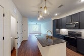 This single-story unit showcases a thoughtfully designed open-concept layout where the kitchen flows seamlessly into the living area. Rich hardwood-style flooring extends throughout the main living spaces, creating a warm and cohesive aesthetic.

The kitchen serves as a central hub, anchored by a white island with dark granite countertops and an integrated single-basin sink with a gold-finished faucet. Overhead, a cluster of glass pendant lights with brass hardware adds a sophisticated touch. Dark navy shaker-style cabinetry lines the walls, extending to the ceiling for ample storage, while a herringbone-patterned tile backsplash and stainless steel Whirlpool appliances complete the functional yet stylish space.

The living area is bright and open, featuring a ceiling fan and a large window that brings in generous natural light. An additional room, finished with plush carpet, a ceiling fan, and French doors leading to a balcony or patio, offers flexible use as a bedroom or sitting room.

The bathroom features large-format light tile flooring, a dark vanity with granite countertops and gold-accented hardware, a tub-shower combination with tile surround, and a recessed built-in shelving unit, providing both style and practicality.