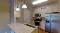 This single-floor residence offers a thoughtfully designed living space centered around a standout kitchen. The kitchen is finished with light granite countertops that extend across a generous island, complete with a double basin sink and pendant lighting overhead — creating both a functional workspace and a natural gathering point. White raised-panel cabinetry lines the walls from counter to ceiling, accented by crown molding details that lend a classic, refined character to the space. Stainless steel appliances, including a range with electric cooktop, an over-the-range microwave, and a full-size refrigerator, provide a modern, polished contrast against the bright cabinetry. Warm hardwood flooring runs throughout, adding depth and continuity to the overall aesthetic. Recessed and overhead lighting ensures the kitchen is well-lit at all times of day. Crown molding details carry through the space, reinforcing the consistent attention to architectural finish. The layout is practical and efficient, with ample storage and counter space to support everyday living. Overall, the space presents a clean, classic interior with quality finishes that balance comfort and style.