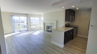 This well-appointed single-floor residence showcases a thoughtfully designed open-concept layout that seamlessly connects the kitchen and living areas. Light hardwood flooring flows throughout, complementing the neutral, crisp white walls and creating a warm, cohesive aesthetic.

The kitchen is equipped with dark espresso cabinetry, stainless steel appliances, and a light-toned granite countertop peninsula that serves as both a prep space and a casual dining area. The peninsula faces the living room, making it ideal for entertaining.

The living area is anchored by a classic white-surround fireplace with a sleek dark insert, providing an elegant focal point. Large corner windows and a sliding glass door flood the space with an abundance of natural light while framing striking panoramic views of the surrounding mountains and landscape. A baseboard heating system is also visible beneath the windows, ensuring year-round comfort.

Recessed lighting in the ceiling adds a modern touch, enhancing the bright, airy atmosphere of the space. The overall ambiance is clean, contemporary, and inviting, combining practical design elements with scenic natural surroundings to create a comfortable and visually appealing living environment.
