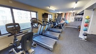 This fitness center is a dedicated, single-floor exercise space designed to support a productive workout routine. The room is outfitted with a row of Cybex-branded treadmills and elliptical machines, all positioned to take advantage of the large windows that line one wall, offering natural light and views of the surrounding exterior. The flooring throughout is a durable, low-pile grey carpet suited for a gym environment. Overhead, recessed track lighting provides consistent, even illumination across the space. A wall-mounted flat-screen television is positioned at the front of the room, adding an entertainment option for those using the cardio equipment. At the far end of the room, a vibrant, large-scale motivational mural depicting an athlete in motion adds energy and visual interest to the space. Additional fitness accessories, including exercise balls, are visible near the mural, suggesting space for supplementary workouts beyond the cardio machines. The overall ambiance is clean, functional, and purpose-driven, making it a practical and inviting environment for daily exercise.