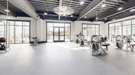 This Nashville fitness center delivers a comprehensive workout environment designed with both functionality and aesthetics in mind. The single-floor layout is organized into distinct training zones, accommodating a wide range of exercise disciplines. The industrial-style design is immediately apparent through the exposed black ceiling structure, visible ductwork, and recessed lighting that illuminates the entire space evenly.

The strength training area is outfitted with an impressive selection of Hoist-brand resistance machines, cable systems, TRX suspension trainers, free weights, and adjustable benches, providing options for targeted muscle training and functional movement. A dedicated cardio zone houses stair climbers, rowing machines, and stationary bikes positioned near large floor-to-ceiling windows that frame views of the surrounding stone-clad exterior courtyard.

A separate open studio area features smooth flooring, ceiling fans, and direct access to an outdoor terrace through full-glass double doors, making it well-suited for group classes or stretching. Throughout the facility, oversized dark-framed windows flood the space with natural light, contributing to an open, energizing atmosphere. Durable rubber flooring provides safety and comfort across all training areas. Full-length mirrors enhance spatial depth and support proper form during workouts.