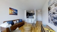 This single-floor space showcases a well-designed open-concept layout that connects the living area and kitchen in a cohesive, functional arrangement. Light hardwood flooring runs throughout, lending warmth and continuity to the space. The living area is comfortably sized, accommodating a leather sofa, a woven ottoman coffee table, and a butterfly accent chair, creating a relaxed yet stylish seating arrangement. A vibrant coastal artwork adds a pop of color to the otherwise neutral, white-painted walls.

The kitchen is positioned toward the rear of the open space and features dark cabinetry that contrasts cleanly against light countertops, stainless steel appliances including a refrigerator, microwave, and range, and a peninsula counter with bar-height seating. This breakfast bar serves as both a functional dining option and a visual divider between the kitchen and living areas.

Recessed and flush-mount ceiling lighting provides even illumination throughout. A hallway visible from the main living area leads to additional rooms, and a white paneled door adds a clean, finished look to the space overall.