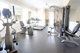 Located at 4129 Inverness Road in Virginia Beach, Virginia, this fitness center offers a clean, well-appointed space designed to support a comprehensive workout routine. The room features durable rubber flooring throughout, providing a safe and functional surface for all types of exercise. Crown molding and wainscoting lend a refined, polished character to the space, elevating it beyond a standard gym aesthetic.

Natural light pours in through multiple large windows dressed with plantation shutters, creating a bright and welcoming atmosphere. Ceiling fans and recessed lighting ensure comfort and visibility at all times of day. A large wall-mounted mirror expands the sense of space and allows for form-checking during workouts.

The fitness center is well-stocked with a variety of cardio equipment, including treadmills, an elliptical trainer, and a recumbent stationary bike. Strength training is supported by a multi-station cable machine, a free-weight rack, and an adjustable weight bench. Additional amenities include a wall-mounted flat-screen television, a water cooler, and a stability ball for functional fitness. Potted plants add a touch of warmth and life to the environment. The space is thoughtfully laid out to accommodate multiple users simultaneously while maintaining an open, uncluttered feel.