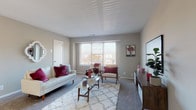 This single-floor residence offers a functional and comfortable layout across several well-defined living spaces. The living room is generously proportioned, featuring neutral gray walls, plush carpeting, and a large window that allows ample natural light to fill the room, creating a bright and welcoming atmosphere.

The kitchen is efficiently designed in an L-shaped configuration, showcasing rich dark mahogany cabinetry accented with gold hardware and light countertops. A full suite of stainless steel appliances includes a gas range, built-in microwave, dishwasher, and refrigerator. Light wood-tone flooring completes the space, providing a clean contrast to the darker cabinetry.

The bedrooms are carpeted and neutral in tone, with windows offering outdoor views of surrounding greenery. Each room provides adequate space for standard furnishings, with closet access noted in at least one bedroom.

The full bathroom features tile flooring, a tiled tub and shower surround, a white vanity with an under-mount sink, and a mirrored medicine cabinet with built-in shelving. A dedicated utility area houses the water heater, adding a practical element to the home's infrastructure.