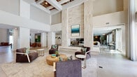 This elegantly designed single-floor amenity space makes an immediate impression with its soaring double-height ceilings, anchored by a dramatic floor-to-ceiling stone-clad fireplace that serves as the architectural centerpiece of the main lounge. The grand proportions of the space are enhanced by large-format tile flooring, white coffered ceiling details with warm wood accents, and an abundance of natural light streaming through tall, floor-to-ceiling windows dressed with sheer drapery. The main lounge area is thoughtfully furnished with plush seating arrangements, including contemporary accent chairs with gold-toned metal legs, organic wood coffee tables, and layered area rugs that define intimate conversation zones. Adjacent to the main area, a cozy lounge features a wall-mounted flat-screen TV, a sectional sofa, and a two-sided fireplace that connects seamlessly to the central gathering space. A dedicated games room offers wood-look tile flooring, a poker table, casual seating, and ample natural light from French doors. A bar-style beverage station with teal counter seating and a private meeting room with glass partitions provide additional functionality. Decorative wall art, billiard cue displays, and curated greenery complete the polished, resort-inspired ambiance throughout.