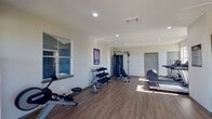 This fitness center is a comprehensively outfitted single-floor facility designed to support a wide range of workout routines. The space features wood-look flooring throughout, recessed lighting, and multiple windows that invite generous natural light and views of the surrounding outdoor environment, creating a bright and motivating atmosphere.

The layout is organized to accommodate various exercise disciplines. A dedicated cardio zone houses treadmills, an elliptical, and a rowing machine, positioned to take advantage of natural light from nearby windows. Strength training is well-represented with a multi-station cable machine, an adjustable weight bench, and a full three-tier dumbbell rack stocked with a varied selection of free weights. A wall-mounted exercise chart provides workout reference for users.

A separate area of the facility is devoted to functional and flexibility training, featuring a vertical rack of medicine balls in multiple weights alongside large stability balls in assorted sizes. A stationary cycling bike adds another cardio option in this zone. A wall-mounted sign displays the fitness center rules and emergency contact information, indicating this is a shared community facility. A mini-split HVAC unit ensures climate control, while the open floor plan allows for comfortable movement between equipment throughout the space.