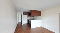 This single-floor space offers a practical and cohesive open-concept layout, where the kitchen and living area flow naturally into one another. The kitchen is anchored by rich, dark-toned wood cabinetry that provides generous storage both above and below the counter. A peninsula with a white base extends from the kitchen, creating a natural divide between the cooking and living spaces while offering additional counter space. The countertops feature a speckled granite-style finish, complementing the deep tones of the cabinetry. A sink sits neatly along the back wall beneath the upper cabinets, and appliances are integrated into the layout for a streamlined look. A textured popcorn ceiling runs throughout, adding a classic character to the interior. Warm hardwood-style flooring extends across the entire space, lending a cohesive and inviting feel underfoot. Neutral beige walls keep the space bright and versatile. A hallway visible from the main area leads to additional spaces within the unit, including what appears to be a closet. The overall ambiance is clean, functional, and move-in ready, suited for everyday living.