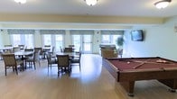 This single-floor community amenity space is thoughtfully designed to encourage social interaction and leisure. The open-plan layout seamlessly integrates multiple activity zones, creating a versatile and inviting environment.

At the heart of the space is a full-size billiards table with a rich burgundy felt surface, serving as a central focal point for recreation. Adjacent to it, a comfortable lounge area is furnished with upholstered sofas and armchairs, complemented by a wall-mounted flat-screen television, making it an ideal spot for relaxation and casual gatherings.

A dedicated dining area features multiple round tables and cushioned chairs, providing ample seating for group meals or social events. Large, multi-pane windows dressed with patterned valances line the walls and flood the room with generous natural light, creating a bright and airy atmosphere throughout the day. A glass-paneled door offers direct access to the outdoors.

The space is finished with warm hardwood flooring and soft yellow walls accented by white crown molding, contributing to its cheerful and welcoming ambiance. Overhead lighting and decorative greenery add warmth and life to the overall aesthetic, making this a well-rounded and functional communal retreat.