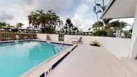 This outdoor amenity space centers around a generously sized swimming pool accented with classic deep blue tile edging, offering clearly marked depth indicators from shallow to a deeper end. The pool's crystal-clear turquoise water is complemented by a broad, light-colored concrete sun deck that surrounds the pool on multiple sides, providing ample room for lounging. Multiple sling-style chaise lounges are positioned throughout the deck, offering residents comfortable spots to relax and enjoy the open sky above.

A distinctive white-painted arched pavilion anchors one side of the pool area, providing a shaded retreat from the Florida sun. This covered structure features smooth stucco finishes consistent with the overall architectural style of the community and includes access to a utility or changing room nearby. Comfortable seating is tucked beneath the archway, creating an inviting shaded gathering spot.

The pool area is enclosed by a white perimeter wall and black iron fencing, ensuring both privacy and security. A vending machine station is conveniently located near one entrance. Surrounding the entire space, tall swaying palm trees and lush tropical greenery create a vibrant, resort-like atmosphere, with views of the broader residential community visible in the background. The overall ambiance is relaxed, tropical, and well-maintained.