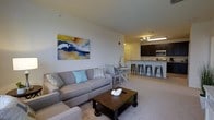 Located at 12001 Old Vine Blvd in Lewes, Delaware, this single-floor residence offers a thoughtfully designed open-concept layout that seamlessly connects the living, dining, and kitchen areas into one cohesive and functional space. The living area is bright and welcoming, with neutral-toned carpeting and ample room for comfortable seating arrangements. A colorful abstract artwork anchors the main wall, adding a vibrant focal point to the space. The kitchen is well-appointed with rich, dark cabinetry, stainless steel appliances including a built-in microwave, and a peninsula with bar-height seating — ideal for casual dining or entertaining. Track lighting illuminates the kitchen area, complementing the sleek countertops and creating a warm, functional atmosphere. Adjacent to the kitchen, a compact dining area provides additional seating with a charming table-and-chair arrangement. The overall design balances practicality with aesthetic appeal, making it well-suited for everyday living. The open flow between spaces enhances the sense of roominess, while the consistent flooring and neutral color palette throughout create a unified, modern look.