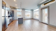 This single-floor unit showcases a thoughtfully designed open-concept layout that seamlessly integrates the kitchen, dining, and living areas. Light hardwood-style flooring flows throughout the main living spaces, contributing to a bright, cohesive aesthetic. Exposed silver ductwork runs along the upper perimeter of the main area, lending an industrial character that contrasts attractively with the otherwise clean, modern interior.

The kitchen is a standout feature, equipped with gray shaker-style cabinetry, a white subway tile backsplash, stainless steel appliances, and a generous center island topped with granite countertops. Cylindrical glass pendant lights hang above the island, adding a refined touch. Large windows along multiple walls of the living area flood the space with natural light and offer views of the surrounding streetscape, while transom windows positioned near the ceiling further enhance the sense of openness and height.

The bedroom is carpeted and includes a well-sized window, maintaining the unit's bright character. The bathroom features light wood-style flooring, a white vanity with a square undermount sink, a large frameless mirror, and a full tub-shower combination. Overall, the space presents a balanced blend of industrial and contemporary design elements.