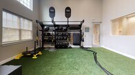 Located near I-75 in Bradenton, Florida, this single-floor fitness facility is designed to support a wide range of training modalities in a clean, purpose-built environment. The space features soaring ceilings that lend an open, airy feel, complemented by an abundance of natural light streaming through arched and rectangular windows positioned at varying heights along the walls.

The training area is divided into distinct functional zones. A large artificial turf surface anchors the center of the space, providing an ideal area for dynamic movement, sled pushes, and battle rope training. A full TRX functional training rig serves as a centerpiece, equipped with suspension trainers, medicine balls in graduated sizes, kettlebells, and a sled. Plyo boxes branded with TRX signage are stacked nearby for plyometric exercises.

A dedicated cardio and conditioning zone features a Concept2 rowing machine and an air assault bike, set on rubber flooring adjacent to the turf. A wall-mounted television and framed artwork contribute to an engaging, motivating atmosphere. Interior pass-through windows add architectural interest while connecting the space visually to adjacent areas. The neutral gray walls and thoughtful equipment organization create a professional, functional environment suited for serious training.