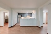 Located at 3155 S Vaughn Way in Aurora, Colorado, this two-bedroom, one-bathroom residence offers a thoughtfully arranged single-floor layout. Light wood-style flooring flows consistently throughout, complementing the clean white walls and dark wood baseboards that define the interior's modern, understated aesthetic.

The combined kitchen and living area serves as the heart of the home. The kitchen features a peninsula with white countertops, slate-blue shaker-style cabinetry extending to the ceiling, stainless steel appliances including a refrigerator and dishwasher, and a range with an overhead vent. The adjacent living area is generously proportioned and open, allowing for flexible furniture arrangements.

Two hallways connect the living spaces to the bedrooms and bathroom. The primary bedroom offers a comfortable footprint with a large window that brings in natural light and outdoor views, plus a built-in wall unit for climate control. The second bedroom also includes a closet with sliding doors. Both rooms maintain the consistent flooring and neutral palette found throughout the home.

The bathroom is well-appointed with a soaking tub and shower combination featuring accessibility grab bars, a curved curtain rod, a gray vanity with white countertop, and a mirrored medicine cabinet. A dedicated closet adds practical storage to round out this efficiently designed residence.