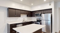This well-appointed 3-bedroom, 1-bathroom unit offers a thoughtfully designed single-floor layout with a clean, contemporary aesthetic throughout. The open-concept kitchen is a standout feature, showcasing rich dark espresso cabinetry, white quartz countertops, a central island, and a full suite of stainless steel appliances including a refrigerator, range, microwave, and dishwasher. Recessed lighting brightens the space, complementing the warm wood-style flooring that extends through the main living and kitchen areas. A wide hallway connects the living spaces to three well-sized bedrooms, each finished with plush carpeting and ceiling fans. The primary bedroom offers generous square footage and access to a walk-in closet with built-in shelving and hanging rods. The full bathroom is finished with wood-look tile flooring, a dark vanity with a white countertop, a large mirror, and a tub-shower combination. A dedicated in-unit laundry room accommodates a full-size washer and dryer. The unit also includes a private balcony with open railings and scenic views of the surrounding landscape and distant mountains, adding an inviting outdoor retreat to this functional and modern living space.