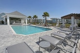 This single-story outdoor pool pavilion offers a welcoming communal retreat designed with both style and function in mind. The structure is defined by striking vaulted ceilings with exposed white beams and green shiplap paneling, creating a bold yet inviting architectural statement. The ceiling's cathedral-like pitch adds a sense of spaciousness and grandeur to the covered area.

The flooring throughout is a classic herringbone-pattern paver, lending a refined, resort-like quality to the space. Outdoor seating is thoughtfully arranged, including a dining table with chairs for alfresco gatherings and additional lounge furniture positioned to take advantage of the open surroundings.

The pavilion's open-air design frames sweeping views of swaying palm trees, a serene community pond, and the pastel-toned residential buildings beyond a wrought iron perimeter fence — creating a bright, tropical ambiance throughout. Abundant natural light fills the space, enhancing its airy and relaxed atmosphere.

Interior access points are visible within the pavilion, suggesting supporting amenities such as restrooms or utility spaces nearby. The combination of crisp white trim, vibrant green accents, and durable outdoor materials makes this pavilion an attractive and functional centerpiece for outdoor community living.