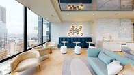 This elegantly appointed high-rise lounge occupies a single floor and immediately impresses with its refined, contemporary design. Expansive floor-to-ceiling windows line the exterior walls, flooding the space with natural light and framing dramatic panoramic views of the surrounding urban skyline. A private balcony is accessible through large glass doors, extending the experience of the cityscape directly into the living environment.

The main lounge area features herringbone-patterned hardwood flooring that runs throughout, adding warmth and visual texture to the modern aesthetic. A sleek marble fireplace serves as a sophisticated focal point, while carefully curated seating arrangements — including teal velvet banquette seating, accent chairs with gold-toned frames, and rounded ottomans — create an inviting, social atmosphere.

An artistic wall installation of metallic spherical pendants doubles as both lighting and sculptural decor, casting a warm ambient glow across the space. Textured wall panels in soft, neutral tones contribute to the overall sense of calm refinement. A dedicated entryway features a frameless glass sliding door and dimensional wall art, providing a striking and gallery-like transition into the main areas.

The open-plan layout encourages flexible use of the space, whether for intimate gatherings or larger social functions. Every detail reflects a high standard of interior design, making this a standout urban retreat.