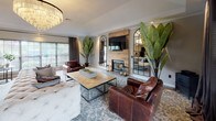 This single-story space is thoughtfully designed, blending modern finishes with warm, inviting elements across its key areas. The living room serves as the centerpiece, anchored by a striking wood-paneled fireplace wall that houses a mounted flat-screen TV and a gas fireplace below. Arched mirror panels flank the fireplace, adding depth and elegance to the room. Rich brown leather armchairs and a tufted sofa surround a herringbone-patterned coffee table set atop a patterned area rug, creating a comfortable and stylish gathering space. Large tropical plants add a lively, natural touch, while French doors and windows allow natural light to filter in, offering glimpses of an outdoor pool area. A statement chandelier completes the polished aesthetic. The kitchen is efficiently laid out in a U-shape, featuring white shaker-style cabinetry, dark granite countertops, and a full suite of stainless steel appliances, including a range, built-in microwave, refrigerator, and dishwasher. A window above the sink frames a direct view of the outdoor pool, enhancing the bright, functional workspace. Wood-look flooring ties both areas together seamlessly.
