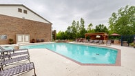 Located on Savannah Circle in O'Fallon, Missouri, this community amenity space offers residents two distinct areas designed for recreation and wellness. The outdoor pool area is a standout feature, showcasing a generously sized swimming pool with brick-accented coping and a wide surrounding deck furnished with lounge chairs for relaxing. A covered wooden pavilion equipped with an outdoor television provides a shaded gathering spot, while colorful landscaping and mature trees frame the area, creating a lush, private atmosphere. A dedicated pool house with classic pitched roofing and multiple French doors connects seamlessly to the pool deck, offering convenient indoor-outdoor access. The second area is a well-appointed fitness center finished with cool blue walls and durable rubber flooring. It is equipped with a comprehensive selection of strength training machines, a cable cross station, free weights with a full dumbbell rack, exercise balls, and cardio equipment including a treadmill. Full-length mirrors along one wall enhance the sense of space and support functional training. Windows allow natural light to filter in, keeping the environment bright and energizing. Together, these two spaces provide a complete lifestyle amenity experience for the surrounding residential community.