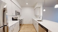 This well-appointed single-floor residence offers a clean, contemporary design throughout, with light wood-style flooring, bright white walls, and abundant natural light creating a fresh and airy ambiance. The galley-style kitchen is a standout feature, showcasing white flat-panel cabinetry with brushed nickel hardware, white quartz countertops, and a full suite of stainless steel appliances including a refrigerator, electric range, microwave, and dishwasher. A stainless steel undermount sink with a modern gooseneck faucet completes the space, while pendant lighting adds a refined touch over the adjoining living and dining area. The open living area benefits from a wall-mounted HVAC unit and a window with outdoor views, bringing in natural light and ventilation. Multiple bedrooms feature plush carpeting, large windows with blinds, and dedicated HVAC vents, offering comfortable and quiet retreats. Two full bathrooms are finished with wood-look flooring, full-size tub/shower combinations with curved curtain rods and grab bars, and clean white fixtures. A dedicated in-unit laundry closet and an electrical panel are conveniently tucked into the hallway, adding practical utility to this thoughtfully designed residence.