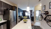 This single-floor residence showcases a thoughtfully designed open-concept layout that seamlessly connects the kitchen and living areas. The kitchen is anchored by a sleek white countertop island with bar seating, providing both workspace and a casual dining option. Stainless steel appliances, including a full-size refrigerator, complement the dark cabinetry, creating a striking contrast. Overhead, a pair of cylindrical pendant lights with chrome fixtures illuminate the island, adding a modern, polished touch.

The living area extends naturally from the kitchen, featuring a bold dark accent wall that creates a dramatic yet inviting atmosphere. A ceiling fan overhead provides both function and style. The space accommodates a comfortable seating arrangement, with natural light filtering in through windows dressed with blinds. Adjacent to the main living area, a partially visible room features a large mirror and striped accent details, suggesting an additional functional space. Light hardwood-style flooring runs throughout, tying the areas together cohesively and enhancing the overall sense of an open, well-appointed interior.
