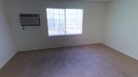This single-floor space presents a straightforward and functional interior. The main room visible is finished with wall-to-wall carpeting in a neutral beige tone, providing a modest and practical foundation underfoot. The walls are painted in a light, neutral color, contributing to a clean and understated aesthetic throughout the space.

A prominent feature of the room is the large window fitted with a metal security grille, allowing a generous amount of natural light to filter in while maintaining added security. The window offers a view of surrounding greenery and neighboring structures, providing a connection to the outdoors. The natural light enhances the openness of the otherwise simply appointed room.

Climate control is addressed by a wall-mounted window air conditioning unit positioned to the left of the window, ensuring comfort during warmer conditions. The room's layout is open and unobstructed, suggesting versatility in how the space can be arranged and utilized.

Overall, the space presents a no-frills, functional environment with essential amenities in place. Its neutral palette and practical features make it a readily adaptable space suitable for a variety of uses.
