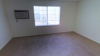 This single-floor space presents a straightforward and functional interior. The main room visible is finished with wall-to-wall carpeting in a neutral beige tone, providing a modest and practical foundation underfoot. The walls are painted in a light, neutral color, contributing to a clean and understated aesthetic throughout the space.

A prominent feature of the room is the large window fitted with a metal security grille, allowing a generous amount of natural light to filter in while maintaining added security. The window offers a view of surrounding greenery and neighboring structures, providing a connection to the outdoors. The natural light enhances the openness of the otherwise simply appointed room.

Climate control is addressed by a wall-mounted window air conditioning unit positioned to the left of the window, ensuring comfort during warmer conditions. The room's layout is open and unobstructed, suggesting versatility in how the space can be arranged and utilized.

Overall, the space presents a no-frills, functional environment with essential amenities in place. Its neutral palette and practical features make it a readily adaptable space suitable for a variety of uses.