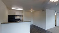 This single-floor space offers a practical and functional layout designed to maximize usability. The open-concept design seamlessly connects the kitchen and living area, creating a cohesive and easy-to-navigate floor plan. The kitchen is equipped with a black gas range, an overhead range hood, and ample white cabinetry that provides generous storage. A peninsula counter with a granite-look surface doubles as a workspace and casual dining area, while a sink adds everyday convenience. The living area is finished with dark hardwood-style flooring that contrasts cleanly against the white walls, lending a contemporary feel to the space. Overhead flush-mount lighting and a fluorescent fixture in the kitchen ensure the interior is well-lit throughout. Ceiling vents provide climate control, contributing to year-round comfort. The clean, neutral color palette across the walls and trim creates a versatile backdrop suitable for a variety of interior styles. The overall ambiance is simple, tidy, and move-in ready, making it an efficient and practical living environment.