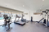 Located at 17301 Keelson Ln in Huntington Beach, California, this well-equipped fitness center offers a clean, modern environment designed to support a full range of workout routines. The single-room facility features smooth concrete-style flooring throughout, crisp white walls, and recessed lighting that creates a bright and energizing atmosphere.

Large floor-to-ceiling glass sliding doors and windows along one wall flood the space with abundant natural light and provide open views of an outdoor pool and patio area, seamlessly connecting the interior to the sunny Southern California surroundings. A wall-mounted split air conditioning unit ensures comfortable temperatures year-round.

The cardio zone is well-appointed with treadmills, an elliptical trainer, and a stationary cycle, all positioned to take advantage of the poolside views. A multi-function cable machine and an adjustable weight bench provide strength training options, while a dedicated free-weight area includes a tiered dumbbell rack stocked with a full range of weights. A large wall mirror in the free-weight section enhances spatial depth and supports proper form during exercise. A wall-mounted flat-screen television adds entertainment to the workout experience. The thoughtful layout ensures ample open floor space for stretching or bodyweight exercises, making this fitness center a functional and inviting resource.