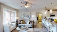This single-floor residence offers a welcoming and thoughtfully designed living area that serves as the heart of the space. The living room is finished with wood-style plank flooring that adds warmth and visual continuity throughout. Neutral wall tones create a clean, versatile backdrop, while natural light filters in through multiple windows dressed with light curtains, brightening the room throughout the day. A ceiling fan with an integrated light fixture is centrally positioned, providing both ambiance and airflow. A large abstract wall painting serves as a focal point above the main seating area, contributing an artistic touch to the otherwise understated palette. The open layout allows for a natural flow from the living area toward adjacent spaces, including a glimpse of a well-lit bathroom featuring warm cabinetry and a mirror — visible through a connecting hallway. The overall design balances comfort and style, incorporating layered lighting through floor lamps and table lamps that complement the natural light. The space presents a calm, airy ambiance well-suited for everyday living.