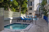 Located at 8665 Burton Way in the heart of Los Angeles, CA, this outdoor amenity space offers a resort-style retreat within a well-maintained residential community. The centerpiece of the area is a generously sized swimming pool with vivid blue tile accents, surrounded by a spacious stone-paved sun deck furnished with lounge chairs — ideal for relaxing under the Southern California sun. Adjacent to the pool sits a separate spa, providing a dedicated space for relaxation and hydrotherapy. The deck is framed by mature trees, including tall magnolias and tropical palms, offering natural shade and a sense of lush seclusion. Bold cobalt-blue planter boxes add a striking pop of color throughout the space, complementing the clean white stucco architecture of the multi-story building above. A separate courtyard patio area features a large dining table with seating for multiple guests, set beneath an open-air atrium with arched windows — perfect for outdoor gatherings. Glass block privacy walls define the perimeter of the pool area while allowing diffused natural light to filter through. The overall ambiance is bright, open, and effortlessly Californian, combining elegant architectural details with inviting, functional outdoor living spaces.