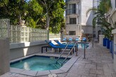 Located at 8665 Burton Way in the heart of Los Angeles, CA, this outdoor amenity space offers a resort-style retreat within a well-maintained residential community. The centerpiece of the area is a generously sized swimming pool with vivid blue tile accents, surrounded by a spacious stone-paved sun deck furnished with lounge chairs — ideal for relaxing under the Southern California sun. Adjacent to the pool sits a separate spa, providing a dedicated space for relaxation and hydrotherapy. The deck is framed by mature trees, including tall magnolias and tropical palms, offering natural shade and a sense of lush seclusion. Bold cobalt-blue planter boxes add a striking pop of color throughout the space, complementing the clean white stucco architecture of the multi-story building above. A separate courtyard patio area features a large dining table with seating for multiple guests, set beneath an open-air atrium with arched windows — perfect for outdoor gatherings. Glass block privacy walls define the perimeter of the pool area while allowing diffused natural light to filter through. The overall ambiance is bright, open, and effortlessly Californian, combining elegant architectural details with inviting, functional outdoor living spaces.