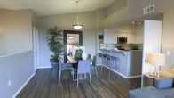 This single-floor residence offers a thoughtfully designed open-concept layout that seamlessly connects the main living areas. The dining space is anchored by a rectangular dark-wood table surrounded by contemporary gray chairs, overhead a sleek pendant light adds a refined focal point. A large framed mirror and lush potted tropical plants bring depth and life to the dining area, enhancing the welcoming ambiance.

Adjacent to the dining space, the kitchen is defined by crisp white cabinetry, stainless steel appliances including a microwave and range, and a peninsula counter with bar-height seating. The peninsula serves as both a functional workspace and a casual gathering spot, offering a natural transition between the kitchen and dining areas.

Rich, dark hardwood-style flooring runs throughout, providing warmth and a cohesive visual flow across the space. The neutral gray wall tones complement the modern furnishings and create a calm, versatile backdrop. Recessed and track lighting in the kitchen, combined with the pendant fixture in the dining area, ensure the space is well-lit and inviting. Overall, the residence strikes a balance between contemporary style and everyday functionality.