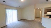 This single-story space presents a clean, functional layout with a comfortable living area as its centerpiece. The main room features neutral-toned carpeting underfoot and light beige walls that contribute to a bright, airy atmosphere. A large sliding glass door with vertical blinds along one wall allows ample natural light to fill the space, creating a welcoming environment throughout the day. Flush-mounted ceiling lighting provides additional illumination when needed.

A distinctive arched passageway connects the carpeted living area to an adjacent entryway or hallway, where the flooring transitions to a light-toned tile in a diagonal pattern. This architectural detail adds a subtle design element while clearly delineating the functional zones of the space. The entryway features a solid door with standard hardware, providing a defined point of entry.

The overall design is straightforward and practical, with smooth walls, standard baseboards, and a ceiling vent for climate control. The neutral color palette throughout makes the space highly adaptable for a variety of furnishing styles and personal touches.
