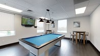 This contemporary game room is designed with both functionality and comfort in mind, offering a well-appointed space for recreation and socializing. The centerpiece of the room is a full-size pool table with a striking blue felt surface, finished in a sleek white frame and illuminated by a classic three-pendant billiard light fixture suspended from above. Dark tile flooring with a wood-look pattern runs throughout, providing a clean contrast to the bright white walls and drop ceiling with recessed panel lighting.

A wall-mounted flat-screen television is prominently positioned for easy viewing from multiple areas of the room, making it ideal for watching sports alongside a game of pool. In a corner nook framed by multiple windows with roller shades, a round high-top table with modern bar stools creates an inviting spot to gather. Natural light filters in from several windows, lending the space an open and airy feel.

The walls are adorned with colorful abstract artwork, adding visual interest and a creative touch to the overall atmosphere. A wall-mounted cue rack keeps billiard equipment neatly organized and accessible. A light wood-framed door with a frosted glass panel provides access to an adjacent area, completing this well-designed, multi-functional recreational space.