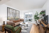 This single-floor residence offers a thoughtfully designed layout that balances functionality with modern aesthetics. The open-concept living area is anchored by light hardwood flooring and features a sliding glass door that leads to a private patio, filling the space with natural light and providing views of the surrounding exterior. Large-scale nature-themed artwork adorns the walls, adding visual warmth to the bright, neutral palette throughout.

The living area accommodates both a comfortable seating arrangement and a dedicated workspace, with a leaning shelf unit and desk tucked along one wall. A round marble-top coffee table and patterned area rug define the central lounge zone, while a full media console completes the space.

The kitchen is clean and contemporary, showcasing white cabinetry, white quartz countertops, and a full suite of stainless steel appliances including a refrigerator, range, and microwave. A peninsula with bar seating and pendant lighting above serves as both a prep surface and casual dining area.

The bedroom is carpeted and calm, featuring a window with slatted blinds that allows soft, diffused light. A circular mirror and nature-inspired wall art contribute to the serene, cohesive aesthetic carried throughout the unit.