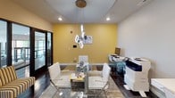 This single-floor space showcases a vibrant, contemporary design anchored by bold yellow accent walls that energize the interior throughout. The main meeting area centers around a sleek glass-top conference table surrounded by modern white chairs, set atop a textured area rug over dark tile flooring. A striking geometric pendant light fixture descends from a curved ceiling detail above, adding architectural interest and focused illumination.

A curated white open-cube shelving unit lines one wall, displaying decorative accessories beneath a series of four coordinating nature-inspired art panels. Adjacent to the conference area, a comfortable lounge nook features gold-and-black striped accent chairs paired with a compact wood side table, offering an inviting informal seating option.

Large floor-to-ceiling glass sliding doors with dark metal frames separate the main area from an adjoining space, allowing natural light to flow freely while maintaining visual connectivity. A dedicated workstation zone accommodates two computer desks and a multifunction printer, making the space fully functional for business operations. The overall ambiance is professional yet welcoming, blending vivid color with polished, modern furnishings.