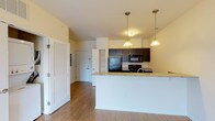 Located in Louisville's vibrant NuLu neighborhood, this single-floor unit offers a well-designed open layout that integrates the kitchen and living areas seamlessly. Light-toned hardwood-style flooring runs throughout the main living spaces, contributing to a clean, contemporary aesthetic.

The kitchen is a functional galley-style space defined by dark espresso cabinetry, granite countertops, and black appliances including a refrigerator, range, microwave, and dishwasher. A peninsula with a granite surface extends into the living area, providing additional workspace and serving as a natural dividing element between the kitchen and the open living space. Pendant lighting suspended above the peninsula adds warmth to the kitchen area.

The living area is generously proportioned, filled with natural light, and opens directly to a private balcony through a sliding glass door, offering views of the surrounding NuLu neighborhood. A dedicated in-unit laundry closet with a stacked washer and dryer is conveniently positioned near the entryway.

The bedroom features plush carpeting and large windows that bring in abundant natural light while providing urban views. The warm, neutral color palette used throughout creates a cohesive and inviting ambiance across the entire space.
