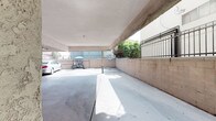 This covered parking area is a practical, ground-level space designed to accommodate multiple vehicles. The structure features a low-profile overhead cover supported by concrete beams, beneath which exposed utility pipes and conduits run along the ceiling, providing functional infrastructure access. The floor throughout is finished in smooth poured concrete, offering a durable and easy-to-maintain surface.

The perimeter of the parking area is defined by sturdy concrete block walls, constructed with a tan-toned masonry finish. Atop these walls, a continuous black wrought iron fence adds a layer of security while allowing visual openness to the surrounding exterior. A gated entry point within the fencing provides controlled access to the property.

The space receives natural light from the open sides of the covered structure, creating a bright environment despite the overhead canopy. Glimpses of surrounding greenery, including trees and shrubs visible beyond the perimeter fence, provide a modest natural backdrop. The layout accommodates both vehicle parking and bicycle storage, with ample room to maneuver. Overall, the parking area is a well-structured, utilitarian space that prioritizes security, accessibility, and efficient use of ground-level square footage.