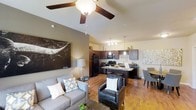 This single-floor residence showcases an open-concept layout that seamlessly connects the living, dining, and kitchen areas into one cohesive and functional space. Vaulted ceilings with a ceiling fan lend a sense of airiness and volume to the living area, which is anchored by a comfortable gray sofa, a leather accent chair, and tasteful wall art that adds a contemporary touch.

The kitchen is a standout feature, boasting rich dark wood cabinetry, granite-look countertops, and a peninsula-style island that doubles as a breakfast bar. Black appliances, including a full-size refrigerator, stove, and microwave, complement the dark cabinetry, while pendant lighting above the island adds warmth and visual interest.

Warm hardwood-style flooring runs throughout the main living areas, tying the open floor plan together and enhancing the welcoming ambiance. A dining area is nestled nearby, providing a natural transition between the kitchen and living spaces. Recessed and pendant lighting fixtures are thoughtfully placed to ensure the space is well-lit throughout. The overall design balances modern aesthetics with everyday practicality, creating an inviting atmosphere suited for both relaxing and entertaining.
