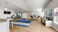 Located at 5003 Turnbridge Cir in Greensboro, NC, this single-story community space offers a thoughtfully designed, open-concept layout that seamlessly integrates a fully equipped kitchen with a comfortable living and gathering area. The soaring vaulted ceilings lend an airy, expansive feel throughout, while large windows fitted with white blinds invite generous amounts of natural light across the light-toned hardwood floors.

The kitchen is outfitted with crisp white cabinetry, granite-style countertops, black appliances, a double-basin sink, and elegant pendant lighting. A peninsula with bar-height seating serves as a natural transition into the social areas of the space, making it ideal for serving and entertaining.

The living area is generously proportioned and versatile, featuring a workstation with dual monitors suited for business use, built-in wall-mounted shelving stocked with media and reading materials, and a seating nook with plush blue accent chairs. A bold, decorative area rug anchors the central floor space, adding a pop of color and visual interest.

Glass-paneled entry doors and multiple windows ensure a bright, welcoming atmosphere, while recessed lighting provides consistent illumination throughout. The clean, neutral palette and cohesive design elements create a functional yet inviting environment well-suited for community gatherings and everyday use.