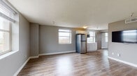 This single-floor unit offers a thoughtfully designed layout that balances open communal areas with defined private spaces. The living area is generously sized, featuring smooth hardwood-style flooring, neutral gray walls, and a textured ceiling. Multiple windows allow natural light to fill the space, creating a bright and welcoming atmosphere. A wall-mounted flat-screen television adds a modern touch to the main living space.

The kitchen is seamlessly integrated into the open-plan area and is equipped with stainless steel appliances, including a full-size refrigerator, a built-in microwave, and a cooktop. White shaker-style cabinetry with gold hardware and light countertops provide ample storage and workspace, while a window above the counter brings in additional natural light.

The bedroom is carpeted with a patterned tile-style carpet, offering a softer, quieter environment. It includes a wall-mounted television, a storage cabinet, a window with exterior views, and a thermostat for individual climate control. An additional room with hardwood-style flooring and a large window provides flexible space for various uses.

The bathroom is finished with wood-style flooring, a gray vanity with a white countertop, a bathtub and shower combination, and chrome fixtures throughout, delivering a clean and functional design.