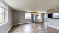 This single-floor unit offers a thoughtfully designed layout that balances open communal areas with defined private spaces. The living area is generously sized, featuring smooth hardwood-style flooring, neutral gray walls, and a textured ceiling. Multiple windows allow natural light to fill the space, creating a bright and welcoming atmosphere. A wall-mounted flat-screen television adds a modern touch to the main living space.

The kitchen is seamlessly integrated into the open-plan area and is equipped with stainless steel appliances, including a full-size refrigerator, a built-in microwave, and a cooktop. White shaker-style cabinetry with gold hardware and light countertops provide ample storage and workspace, while a window above the counter brings in additional natural light.

The bedroom is carpeted with a patterned tile-style carpet, offering a softer, quieter environment. It includes a wall-mounted television, a storage cabinet, a window with exterior views, and a thermostat for individual climate control. An additional room with hardwood-style flooring and a large window provides flexible space for various uses.

The bathroom is finished with wood-style flooring, a gray vanity with a white countertop, a bathtub and shower combination, and chrome fixtures throughout, delivering a clean and functional design.