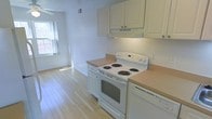 This single-floor space offers a clean, functional layout designed for comfortable everyday living. The kitchen is a standout feature, showcasing white cabinetry that extends to the upper walls for ample storage, paired with neutral-toned countertops that provide generous prep space. White appliances, including a coil-burner electric range with oven and a dishwasher, are seamlessly integrated into the cabinetry for a cohesive, streamlined look. A built-in range hood sits above the stove, adding a practical touch to the cooking area. Light hardwood flooring runs throughout, lending warmth and continuity to the space while reflecting natural light effectively. A window positioned near the kitchen allows natural light to filter in, brightening the interior and creating an open, airy atmosphere. The neutral white and beige color palette throughout keeps the space feeling fresh and versatile. Visible ceiling ventilation ensures consistent airflow, contributing to a comfortable indoor environment. The overall design is straightforward and practical, making efficient use of the available space while maintaining a tidy, welcoming ambiance suited to a variety of occupants.