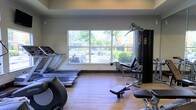 This fitness room offers a practical and well-organized space designed to support a range of exercise routines. The room features hardwood-style flooring throughout, providing a clean and durable surface suited for both cardio and strength training activities. A large picture window dominates one wall, allowing ample natural light to fill the space while offering views of a landscaped outdoor area, complete with greenery, a pergola structure, and open walkways — creating an pleasant atmosphere during workouts.

The equipment selection is comprehensive, including multiple treadmills and a stationary bike for cardiovascular training, alongside a multi-station cable and weight machine for strength exercises. A padded workout bench is also present, adding versatility for free-weight and resistance exercises. The layout is thoughtfully arranged to allow sufficient circulation space between machines.

The room's neutral wall tones and bright natural lighting contribute to an open, airy feel. The single-level layout ensures easy accessibility throughout. Overall, this fitness space is well-suited for residents or guests looking for a convenient and functional workout environment.