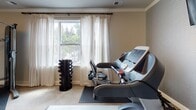 The Bound Brook Fitness Center at 675 Tea St in Bound Brook, NJ, is a thoughtfully appointed single-floor facility designed to support a comprehensive workout experience. The space is finished with neutral-toned walls, crown molding, and plush carpeting, creating an inviting atmosphere that balances functionality with refined aesthetics.

The fitness area is equipped with a treadmill, an elliptical trainer, and a stationary bike, all positioned to maximize the use of available space. A multi-station cable weight machine provides strength training options, while a tower dumbbell rack offers additional free weight exercises. Protective floor mats are placed beneath the equipment to ensure safety and preserve the carpeted flooring.

A wall-mounted flat-screen television provides entertainment during workouts. Elegant drum-shade flush-mount ceiling fixtures with warm gold accents illuminate the space with a soft, welcoming glow. A floor-to-ceiling mirror on one wall expands the visual depth of the room and allows users to monitor their form during exercise.

Natural light filters in through windows dressed with sheer, patterned curtains, offering views of the surrounding greenery. French doors with glass panes serve as the main entry, lending the space a polished, upscale character. The overall ambiance is clean, comfortable, and well-maintained, making it a practical and pleasant environment for daily fitness routines.