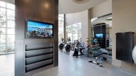 This contemporary fitness center is thoughtfully designed to support a full range of workout routines in a sleek, modern environment. The space is anchored by soaring floor-to-ceiling windows that line the exterior walls, inviting abundant natural light and offering views of the surrounding outdoor areas, including a poolside lounge. The open layout is finished with large-format tile flooring and accented by curved architectural elements and high ceilings that create an airy, spacious atmosphere. A dedicated cardio zone features treadmills, elliptical trainers, and stationary bikes positioned to take advantage of the expansive window views. Strength training is well represented with a lineup of Hoist selectorized machines targeting various muscle groups, complemented by a rack of kettlebells, medicine balls, stability balls, and a full dumbbell rack for functional and free-weight training. Large wall mirrors amplify the sense of space and assist with form, while integrated flat-screen televisions are mounted throughout, including one recessed into a striking dark wood-paneled feature wall with built-in shelving. The fitness center is accessed through full-length glass sliding doors, maintaining visual connectivity with the exterior. Recessed lighting and a refined color palette of dark wood, neutral tones, and polished surfaces give the space a premium, hotel-quality feel.