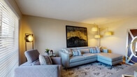 Located at 7218 S Padre Island Drive in Corpus Christi, Texas, this single-floor residence offers a well-curated layout with modern finishes and contemporary décor throughout. The living area serves as the centerpiece of the home, featuring a light gray sectional sofa accented with decorative pillows, a quilted accent chair, and a striking large-scale abstract artwork with bold gold brushstrokes that anchors the room. Warm, layered lighting and a patterned area rug complete the inviting atmosphere, while a window allows natural light to fill the space. The kitchen is outfitted with crisp white cabinetry, granite-look countertops, stainless steel appliances including a gas range, microwave, and dishwasher, and durable wood-look flooring. An adjacent dining area accommodates intimate meals, accented by decorative circular wall art and a bold abstract canvas. The bedroom is a comfortable retreat with plush carpeting, a upholstered headboard, warm wood nightstands, and vibrant framed artwork. A ceiling fan adds practicality, while a dresser and accent chair provide additional functionality. The bathroom features wood-look flooring, a tub-shower combination with white tile surround, a granite-top vanity, and coastal-inspired wall art, creating a clean and relaxing environment.